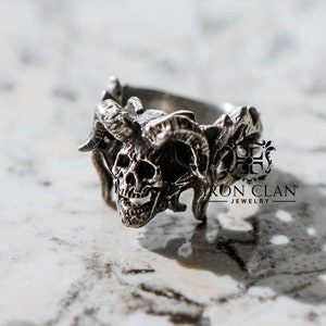Puede incluir: Un anillo de plata con un diseño de calavera y cuernos de carnero. El anillo está hecho por Iron Clan Jewelry.