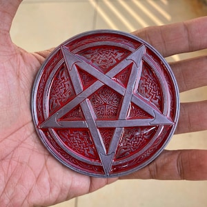 May include: A circular metal medallion featuring a raised silver pentagram centered over a deep red background. The surrounding border and the spaces between the pentagram points are decorated with intricate, embossed Celtic knotwork patterns.