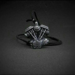 Può includere: Un ciondolo a forma di motore di moto color argento su un cordino nero. Il ciondolo è una miniatura dettagliata di un motore, con cilindri visibili e altri componenti meccanici. Il cordino è nero e sembra essere fatto di un materiale intrecciato. Lo sfondo è grigio scuro.