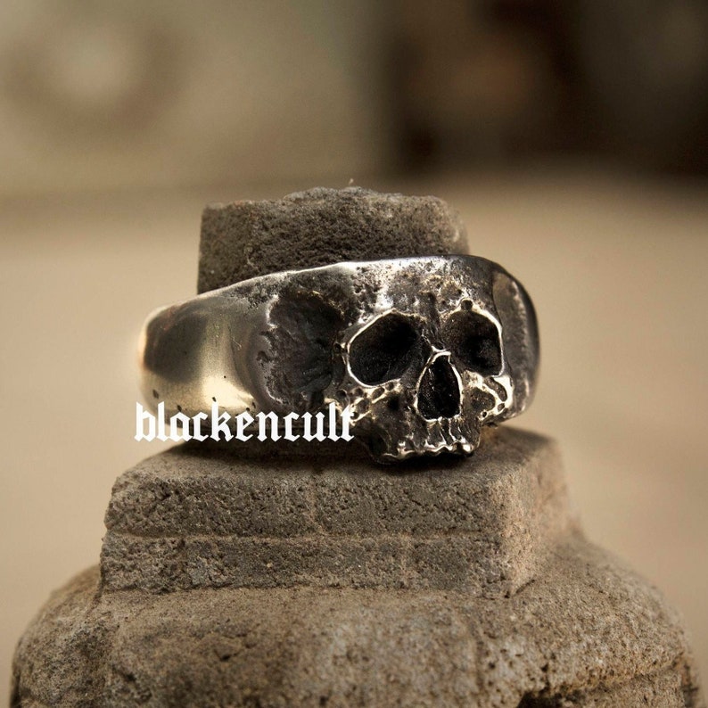 May include: A silver skull ring with a detailed design. The ring is made of metal and has a textured finish.