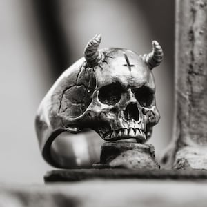 May include: A silver-colored skull ring with horns and a cross symbol on the forehead. The ring has a detailed, textured surface and is set against a blurred background. The skull ring is a gothic-themed accessory.