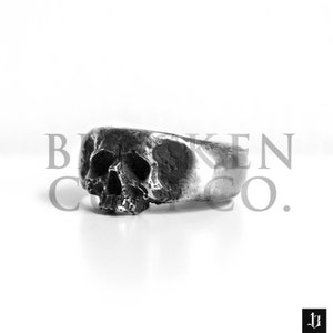 May include: A silver skull ring with a detailed design. The ring is worn on a finger and is a statement piece of jewelry.