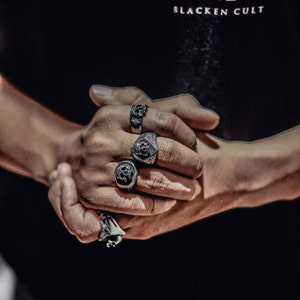May include: A person's hands wearing four silver rings with black skull designs. The rings are worn on the index, middle, and ring fingers.