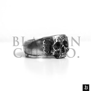 May include: A silver skull ring with a textured finish. The ring is designed to look like a human skull with a detailed face.