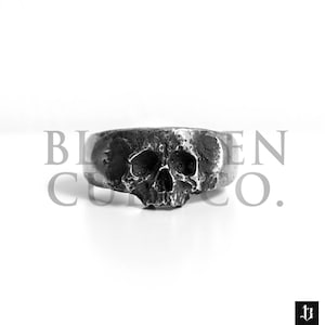 May include: A silver skull ring with a detailed design. The ring is made of metal and has a textured finish.