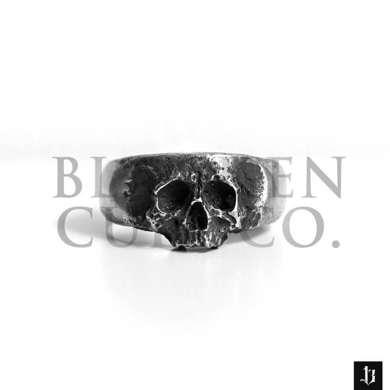 May include: A silver skull ring with a detailed design. The ring is made of metal and has a textured finish.