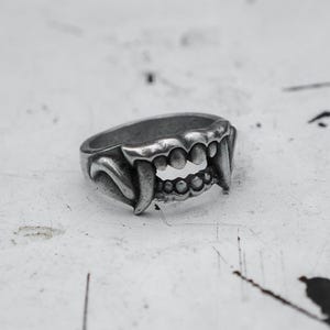 May include: A silver-colored ring with a detailed design of a set of vampire fangs. The ring features sharp, curved canines and multiple smaller teeth, creating a gothic or fantasy aesthetic. The ring is set against a white background.