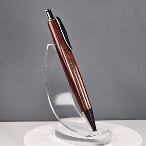Op de afbeelding: Een klik-balpen met een gepolijste donkerbruine houten behuizing, een zilverkleurige metalen drukknop aan de bovenkant, een zwarte zakclip en een taps toelopende zwarte metalen punt, rechtopstaand op een doorzichtige acryl standaard.