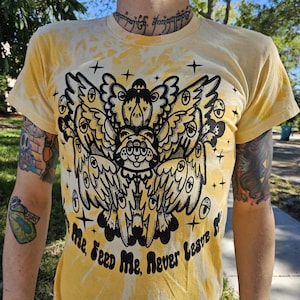 May include: Yellow tie-dye t-shirt with a black and white graphic print of a creature with multiple eyes and wings. The text "Love Me, Feed Me, Never Leave Me" is printed below the graphic.