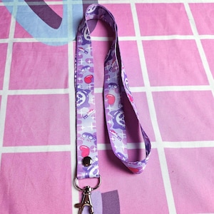 Könnte beinhalten: Ein lilafarbenes Lanyard mit einem sich wiederholenden Muster aus Herzen, Sternen und einer Comicfigur mit einem Messer. Das Lanyard hat einen silbernen Metallclip.
