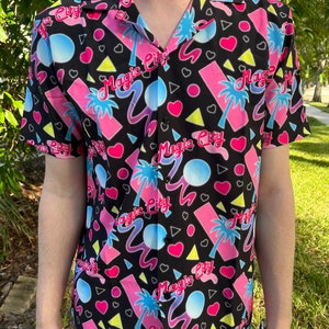 Puede incluir: Una camisa negra de manga corta con botones y un estampado colorido de inspiración retro. El estampado presenta las palabras "Magic City" en una fuente rosa, azul y amarilla, junto con palmeras, corazones y formas geométricas.