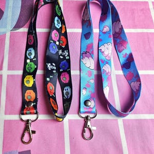 Op de afbeelding: Twee kleurrijke lanyards met zilveren clips. Eén lanyard heeft een zwarte achtergrond met kleurrijke cartoonfiguren. De andere lanyard heeft een blauwe achtergrond met roze en witte cartoonfiguren.
