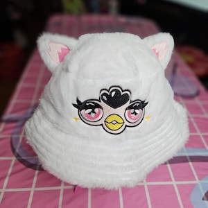 May include: White fuzzy bucket hat with pink ears and an embroidered cartoon bird with pink eyes and a yellow beak.