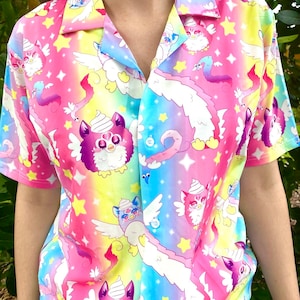 Puede incluir: Una camisa de manga corta con botones, con un fondo degradado de arcoíris y un patrón repetido de criaturas peludas de dibujos animados con alas y estrellas.