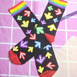 May include: A pair of black socks featuring a pattern of colourful arrows. The socks have red toes and rainbow-striped cuffs. The arrows are in various colours, including red, yellow, green, blue, and purple.