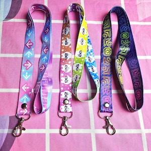 May include: Four colorful lanyards with different patterns and designs. The lanyards have silver metal clips at the end. The lanyards are on a pink and white checkered background.