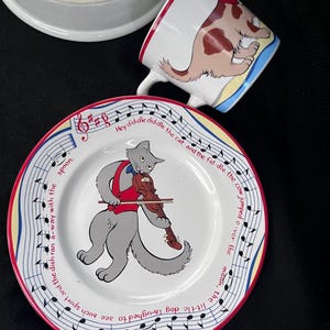 May include: A white ceramic plate with a red rim features a gray cat playing a violin. The plate has a blue and white musical note border with the text "Hey diddle diddle the cat and the fiddle the cow jumped over the moon, the little dog laughed to see such sport and the dish ran a-way with the spoon."