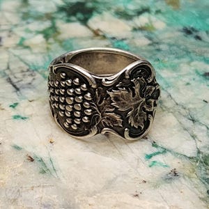 May include: A silver ring with a grapevine design. The ring is made from a repurposed spoon and features a cluster of grapes and a leaf.