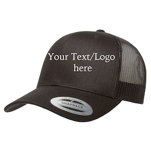 Puede incluir: Gorra de camionero negra y blanca con malla en la parte posterior. La gorra tiene una pegatina plateada en el lateral que dice "The Classics Snapback Yupoong Since 1974". La parte delantera de la gorra tiene el texto "Your Text/Logo here" en blanco.