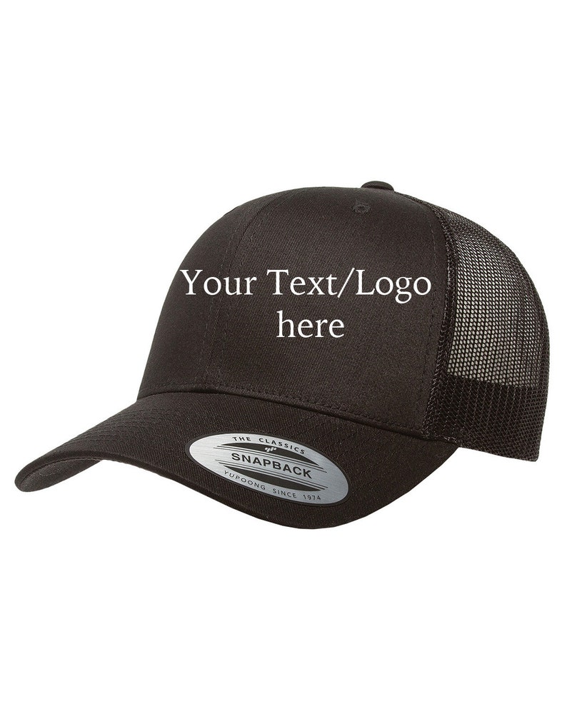 Puede incluir: Gorra de camionero negra y blanca con malla en la parte posterior. La gorra tiene una pegatina plateada en el lateral que dice "The Classics Snapback Yupoong Since 1974". La parte delantera de la gorra tiene el texto "Your Text/Logo here" en blanco.