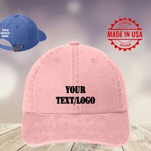 May include: A pink and blue baseball cap with the text "YOUR TEXT/LOGO" embroidered on the front. The cap is made in the USA and is handmade.