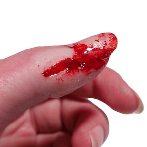 May include: A realistic prosthetic finger tip featuring a simulated injury with bright red, glossy, viscous liquid applied to the surface to mimic the appearance of fresh blood, creating a convincing effect for special effects makeup applications.