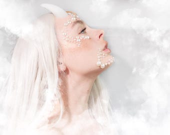 May include: A profile view of a face adorned with white pearl embellishments around the eye and chin, featuring long platinum white hair and a single white horn protruding from the forehead, set against a soft, ethereal cloud-like background.
