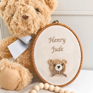 May include: A plush tan teddy bear wearing a white ribbon and a party hat, positioned next to an oval embroidery hoop featuring the name Henry Jude in brown thread and a small crocheted bear face with a brown and white checkered bow.