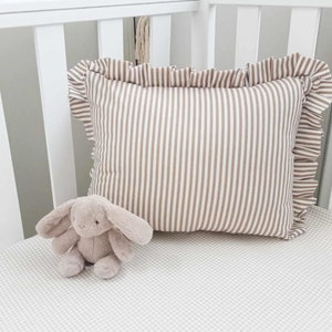 May include: A white cot with a brown and white striped pillow with frills and a grey stuffed bunny on a white and grey gingham cot sheet.