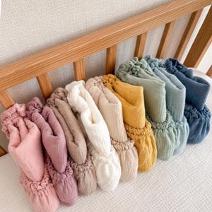 May include: A collection of folded muslin sheets in various colours including light pink, mauve, beige, white, cream, mustard yellow, sage green, light blue, and dark blue, arranged in a row with the text Soft Muslin Sheets at the bottom.
