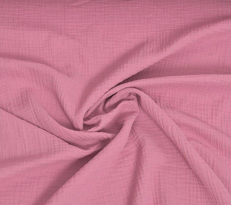May include: A close-up shot of a soft, pink fabric with a textured, crinkled appearance. The fabric is draped and folded, creating shadows and highlights that emphasize its texture. The color is a muted, dusty rose.