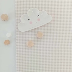 May include: A white ceramic cloud-shaped dish with a pink smiling face and closed eyes. The dish is on a light grey and white gingham fabric background. There are several small wooden and white beads scattered around the dish.