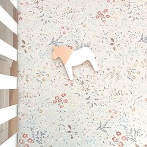 Puede incluir: Un juguete de caballo de madera con cuerpo blanco y cabeza de madera natural descansa sobre una s&aacute;bana con estampado floral. La s&aacute;bana presenta un fondo azul claro con un delicado patr&oacute;n de flores y hojas en tonos apagados.
