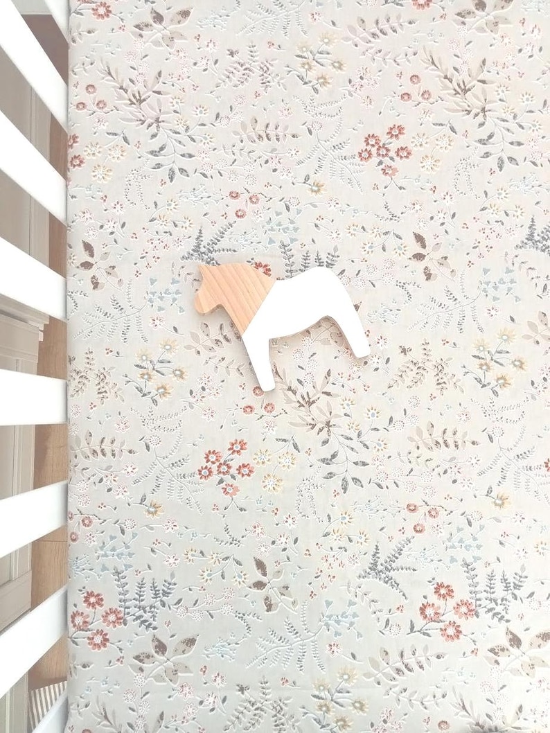 Puede incluir: Un juguete de caballo de madera con cuerpo blanco y cabeza de madera natural descansa sobre una s&aacute;bana con estampado floral. La s&aacute;bana presenta un fondo azul claro con un delicado patr&oacute;n de flores y hojas en tonos apagados.