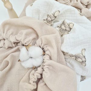 May include: A collection of soft, beige and white baby blankets with a bunny print, arranged in a wicker basket. A sprig of cotton adds a natural touch. The blankets are made of a delicate, textured fabric.