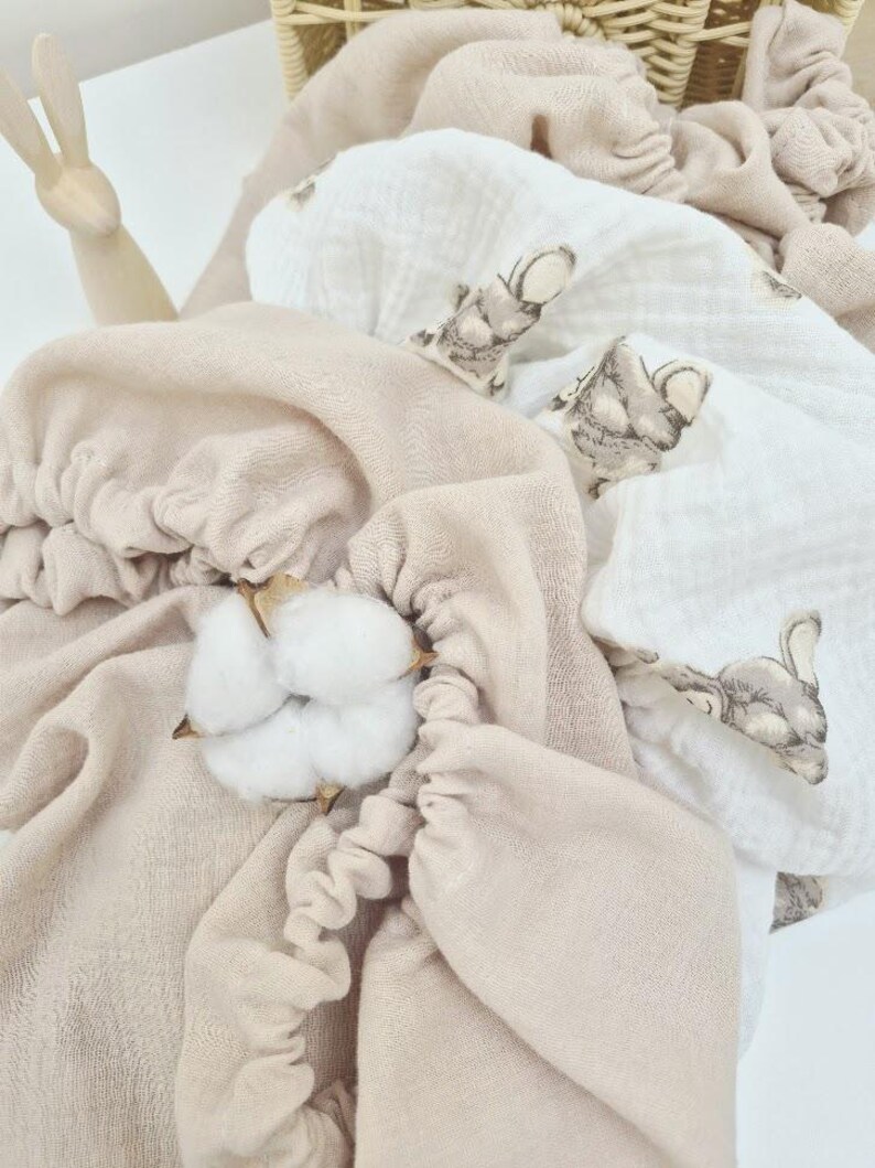 May include: A collection of soft, beige and white baby blankets with a bunny print, arranged in a wicker basket. A sprig of cotton adds a natural touch. The blankets are made of a delicate, textured fabric.