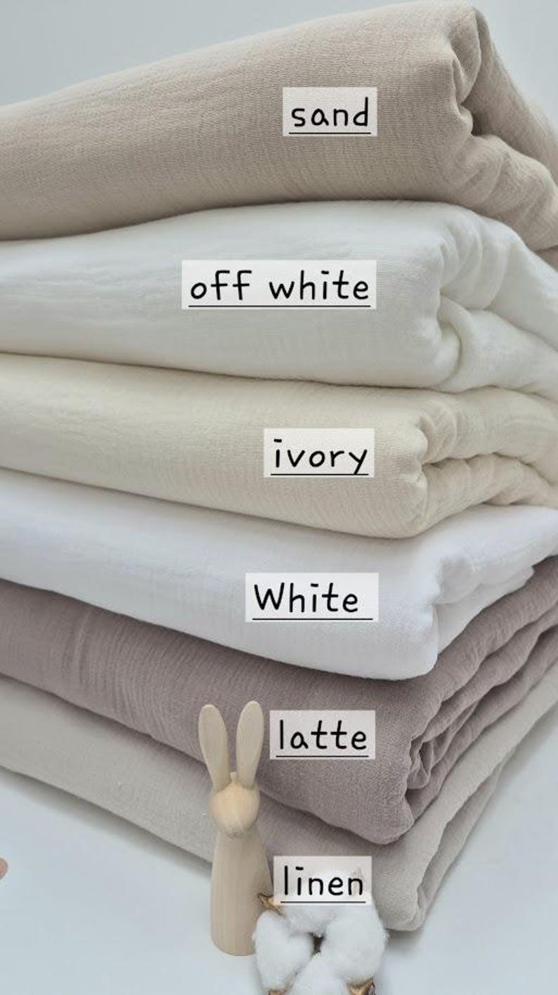 May include: A stack of folded fabric in various neutral colors, including sand, off-white, ivory, white, latte, and linen. A wooden bunny figurine and cotton sprig are in the foreground. The fabric is labeled with its color name.