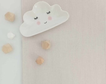 May include: A white ceramic cloud-shaped dish with a smiling face and closed eyes. The dish is on a pink and white striped fabric background. There are four small wooden geometric shapes scattered around the dish.
