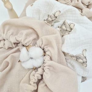 May include: A collection of soft, beige and white baby blankets with a bunny print, arranged in a wicker basket. A sprig of cotton adds a natural touch. The blankets are made of a delicate, textured fabric.