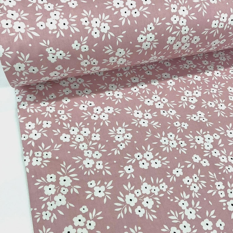 Puede incluir: Un rollo de tela con un estampado floral repetitivo. La tela tiene un fondo rosa empolvado con flores y hojas blancas. Las flores son peque&ntilde;as y agrupadas, creando un dise&ntilde;o delicado y femenino. Ideal para proyectos de costura.