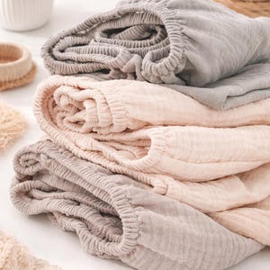 May include: A stack of three folded muslin fabric items with elasticized waistbands. The top item is light gray, the middle item is a soft pale peach, and the bottom item is a muted taupe. Each piece features a crinkled, textured surface.