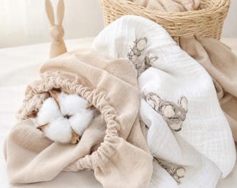 May include: A beige muslin fabric with an elasticated edge holding a white cotton boll, next to a white muslin fabric featuring a repeating grey rabbit print. A small wooden rabbit figurine and a woven wicker basket are visible in the background.
