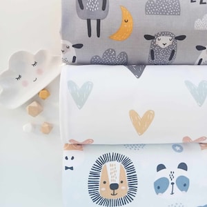May include: Three stacks of patterned fabric with cartoon designs. The top fabric is grey with sheep, clouds, and a crescent moon. The middle fabric is white with blue and gold hearts. The bottom fabric is light blue with a lion and panda.