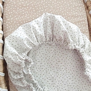 May include: White fitted crib sheet with a delicate floral pattern in light gray. The sheet is gathered around the edges and sits inside a woven bassinet. The background is a light tan fabric with a matching floral print.