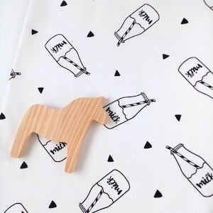 Puede incluir: Un juguete de madera con forma de caballo descansa sobre una tela blanca estampada con ilustraciones de botellas de leche negras y triángulos. Las botellas de leche tienen una pajita y la palabra "milk". El juguete es de madera clara.