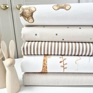 May include: A stack of fabric rolls with various patterns, including teddy bears, stars, stripes, giraffes, and small stripes. Two wooden bunny figurines stand in front of the fabric. The color palette includes white, beige, and brown.