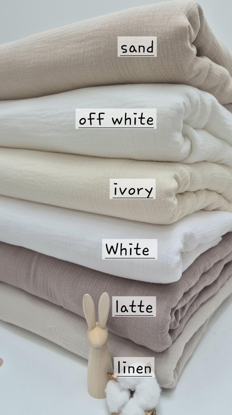 May include: A stack of folded fabric in various neutral colors, including sand, off-white, ivory, white, latte, and linen. Each fabric color is labeled with its name. A small wooden bunny figurine is placed near the bottom of the stack.