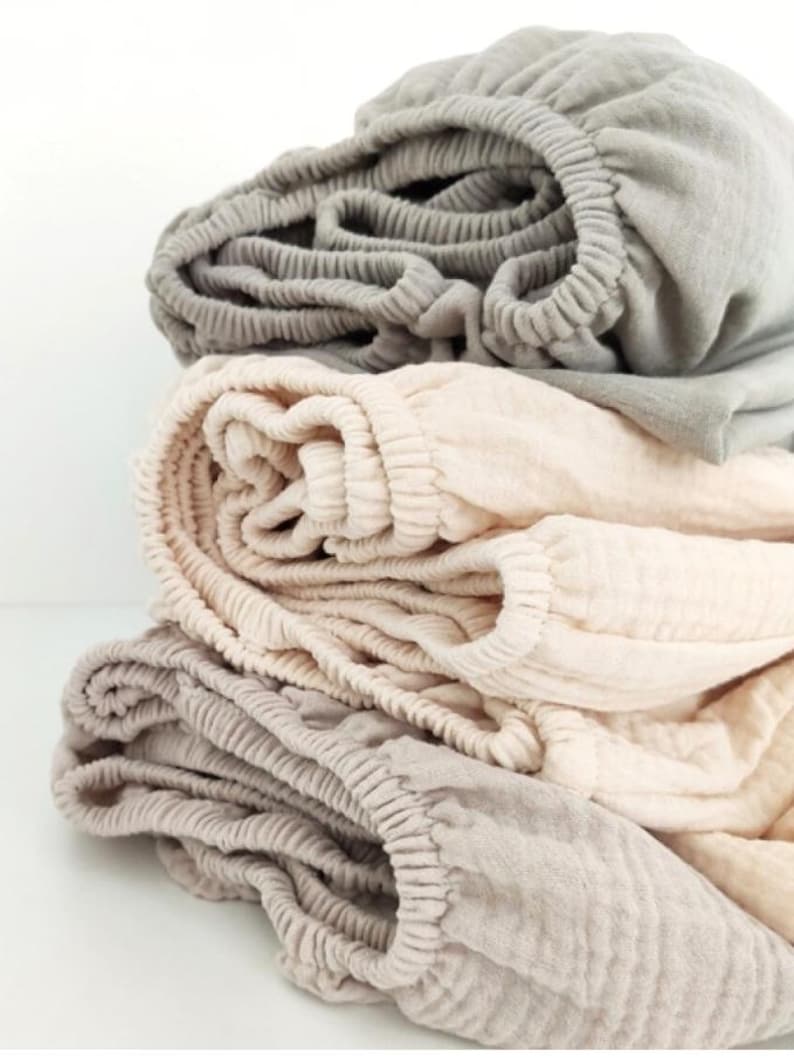 May include: A stack of three fitted bed sheets in neutral colors. The top sheet is a light gray, the middle sheet is a pale peach, and the bottom sheet is a light beige. Each sheet has an elasticized edge.