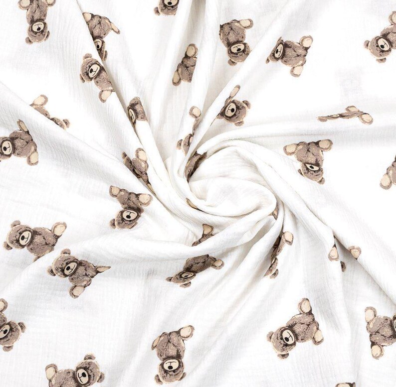 May include: White fabric with a repeating pattern of brown teddy bears. The bears are in various poses, with their heads and limbs visible. The fabric is soft and appears to be a lightweight material, suitable for baby blankets or clothing.