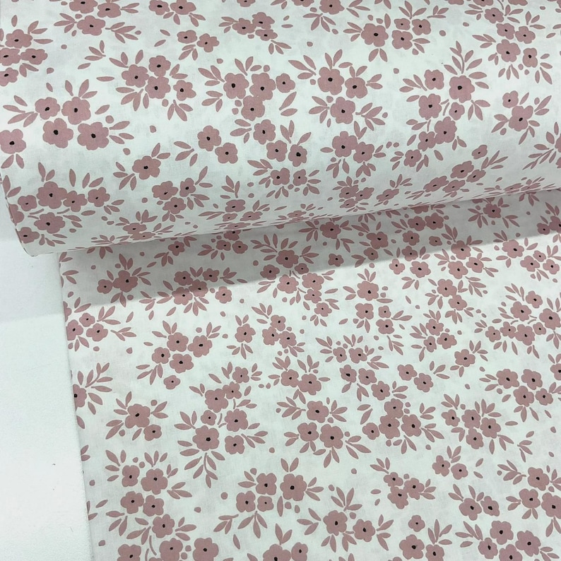 Puede incluir: Rollo de tela blanca con un patr&oacute;n repetido de peque&ntilde;os racimos florales rosas y hojas verdes. Las flores tienen centros oscuros, creando un dise&ntilde;o delicado. La tela es probablemente de algod&oacute;n o similar, adecuada para proyectos de costura o artesan&iacute;a.